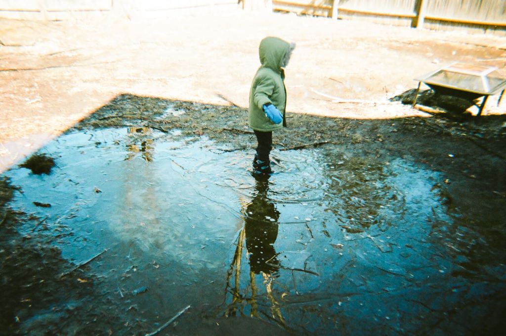 Un profil d'enfant dans un manteau vert avec des mitaines bleues debout dans une flaque d'eau glacée. Le ciel bleu se reflète dans la flaque d'eau. 
 & #10;5 cadres... De Kodak Portra 400 sur un appareil photo plastique Kodak M35 - par Kerry Constantino