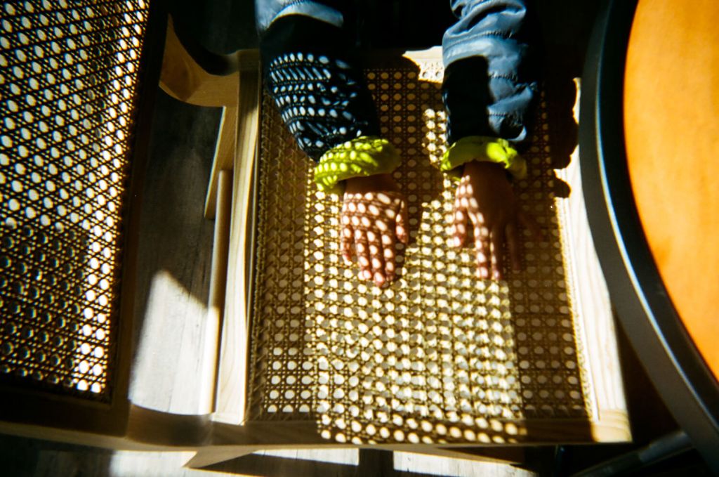 Deux mains reposent sur le siège d'une chaise en osier. Il y a des cercles d'ombre et de lumière sur les mains et les bras. Les mains et les bras portent un manteau bleu marine et jaune vif.
 & #10;5 cadres... De Kodak Portra 400 sur un appareil photo plastique Kodak M35 - par Kerry Constantino