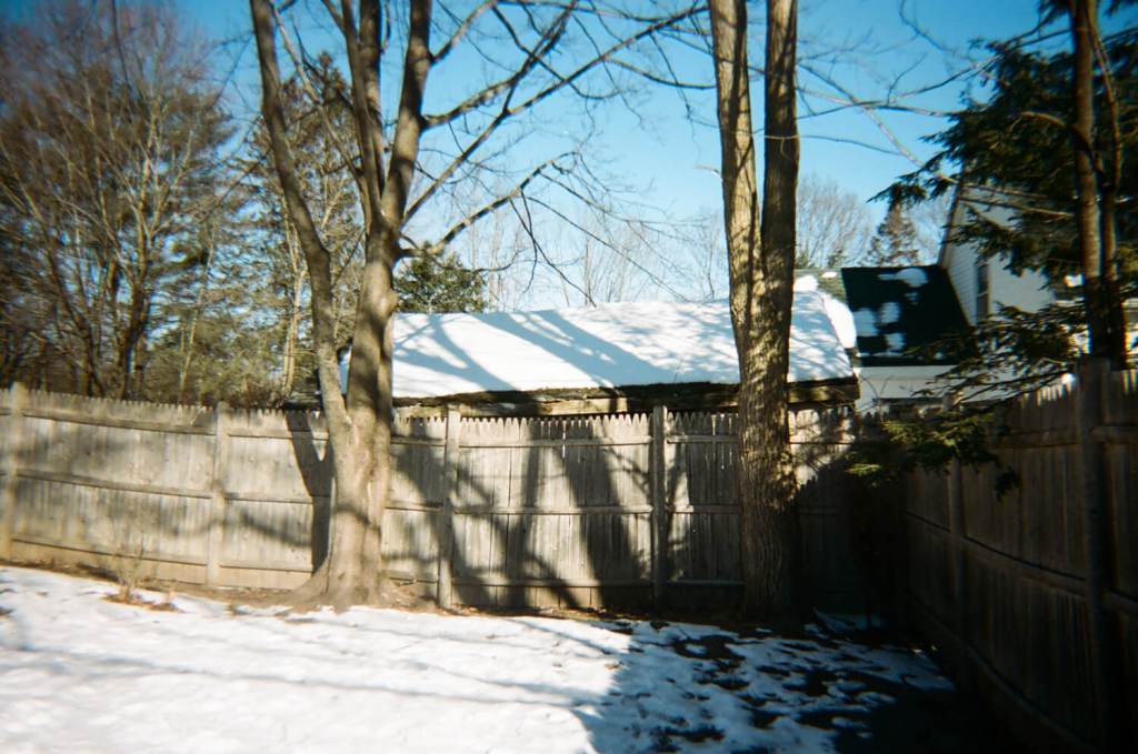 Les ombres des branches d'arbres sont projetées sur une clôture en bois. Il y a de la neige sur le sol derrière la clôture et de la neige sur le toit d'un bâtiment qui se trouve derrière la clôture. Le ciel est bleu. Deux arbres sont devant la clôture.
 & #10;5 cadres... De Kodak Portra 400 sur un appareil photo plastique Kodak M35 - par Kerry Constantino